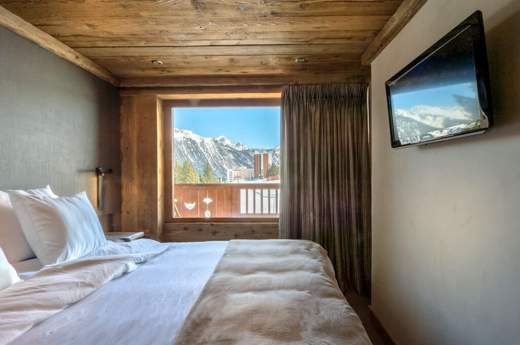 Une chambre à l'esprit cosy dans un appartement à la montagne avec vue sur la piste enneigée. La chambre est décorée d'un mur en tissu et d'un plafond en vieux bois.