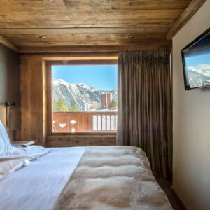 Une chambre à l'esprit cosy dans un appartement à la montagne avec vue sur la piste enneigée. La chambre est décorée d'un mur en tissu et d'un plafond en vieux bois.