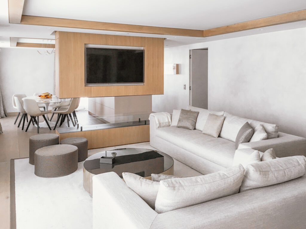 La pièce à vivre dans les tons de beige avec des touches de bois, est divisée par un mur présentant une télévision et une cheminée.